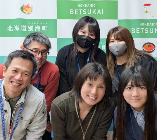 北海道別海町のイベント会場と思われる場所で、6人の男女が笑顔で集合写真を撮っています