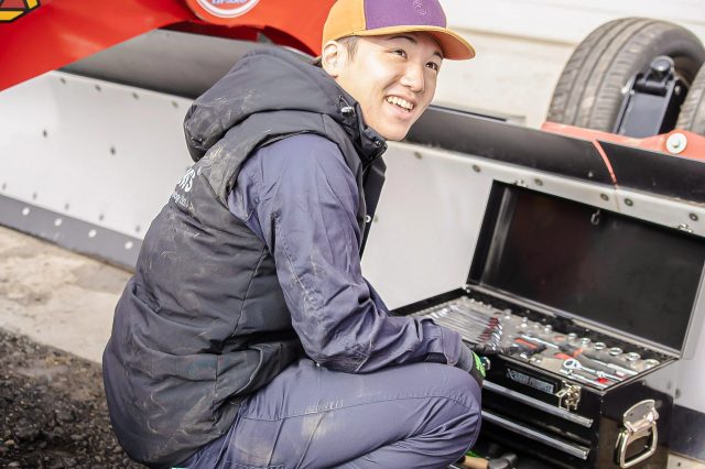 作業服を着た若い男性が、工具箱の前で笑顔を見せています。車両か機械のメンテナンス作業中のようです。