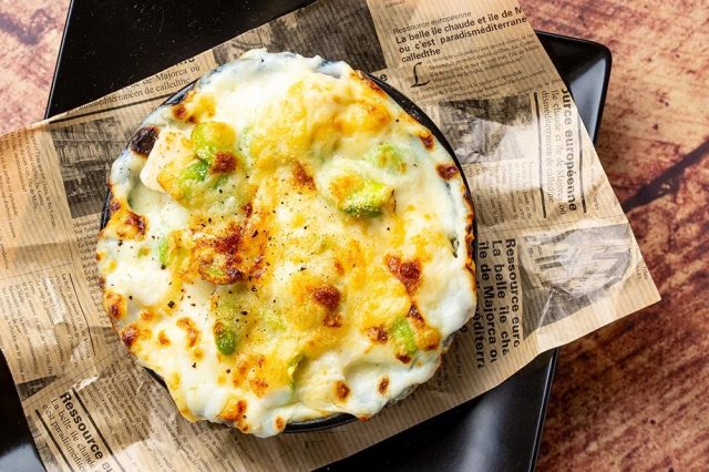 熱々のチーズがたっぷりかかったグラタンのような料理で、緑色の野菜がアクセントになっています